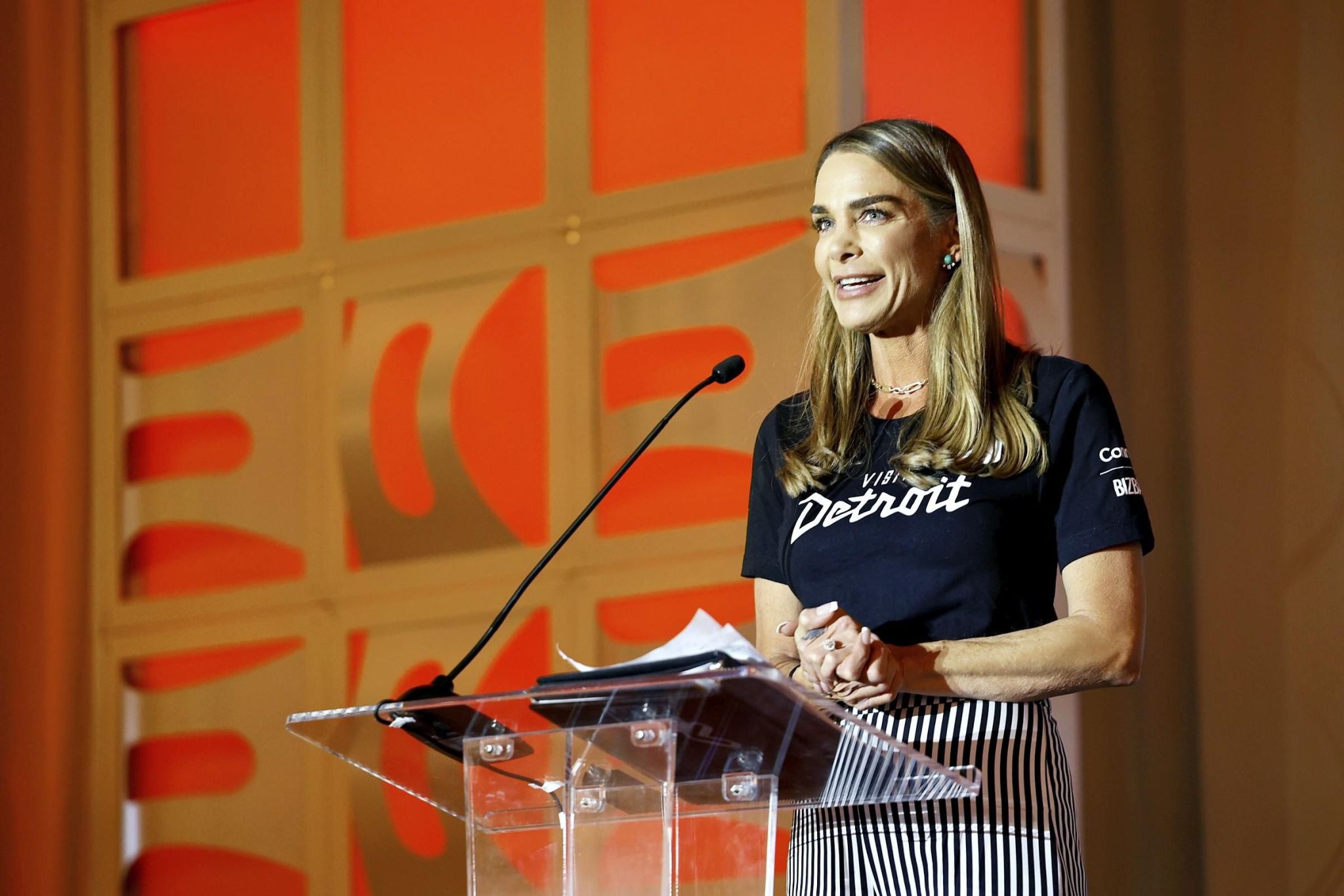 A woman with brown hair wearing a black shirt standing on stage speaking into a microphone on a podium.