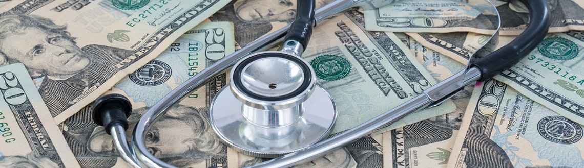 A stethoscope rests on top of scattered U.S. twenty and hundred dollar bills.