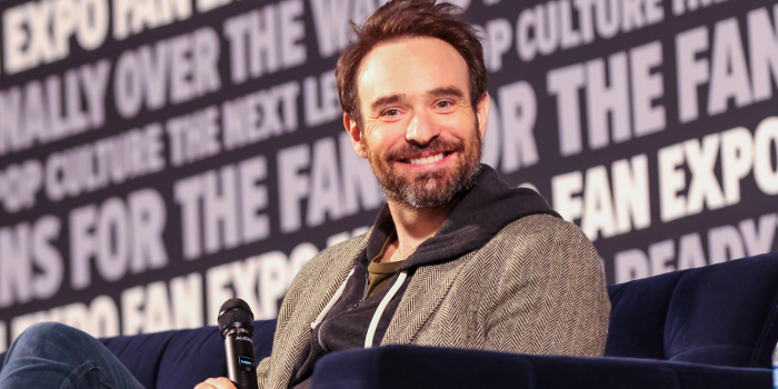 Charlie Cox stares directly into the camera smiling, as he sits on the navy blue couch of the main stage.