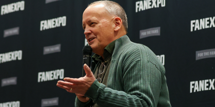 Dee Bradley Baker, in front of the FAN EXPO HQ backdrop, speaks with his hands during his panel at FAN EXPO Portland