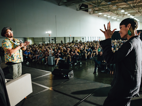 Rosario Dawson walking on stage, smiling and holding her hands up in the ""applause" motion for ASL. There is a large crowd in the background, and a male moderator on the left side of the stage, clapping.