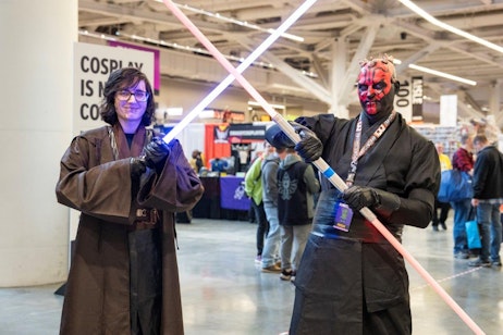 The image captures two individuals in a comic convention setting, dressed in detailed Star Wars-themed cosplay. On the left, a person dressed as Anakin Skywalker, complete with a brown Jedi robe and a blue lightsaber, smiles at the camera. On the right, another cosplayer represents Darth Maul, sporting red and black facial makeup, a black outfit, and wielding a red double-bladed lightsaber. They stand ready in a playful duel pose, adding a dynamic element to the scene, with the bustling convention atmosphere visible in the background.
