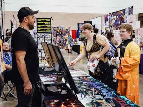 At a bustling convention hall, a vendor in a black cap and t-shirt stands beside his merchandise booth displaying various artwork. He interacts with two enthusiastic attendees: a woman wearing a steampunk-inspired outfit and a young individual clad in an orange costume reminiscent of anime characters. In the background, colorful displays and other convention-goers can be seen, encapsulating the vibrant atmosphere of the event.