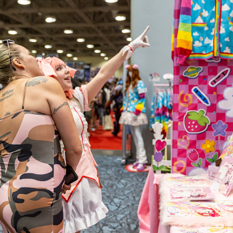 A mother and daughter shop in artist alley. The art at the booth is brightly coloured and whimsical. The young fan points eagerly at an item for sale at the booth. 