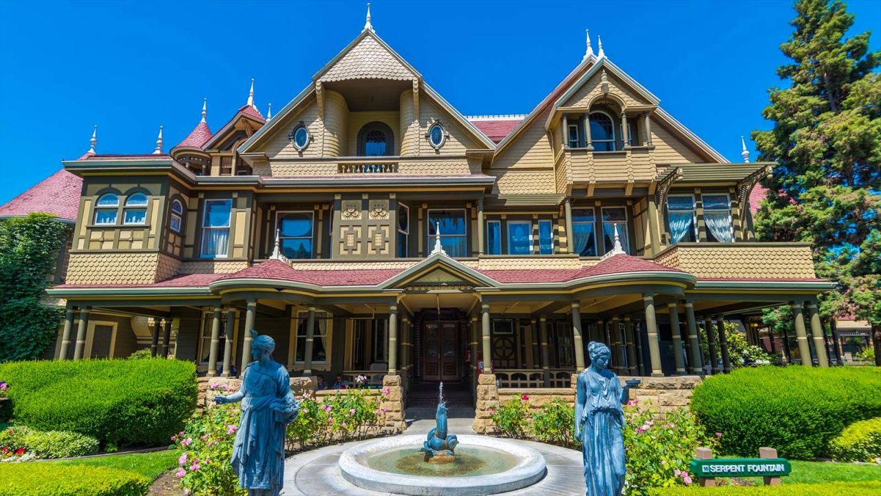 Outside of the mansion, front side, on a beautiful sunny day. Victorian statues stand in front of a serpent statue on the front walk way. This house features many large windows to let the light in and a wrap around porch