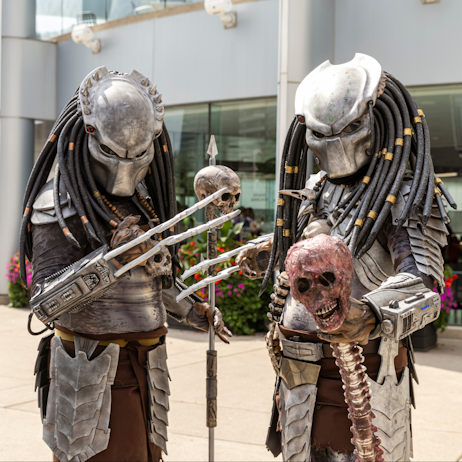 Two cosplayers, both dressed as Predator species, pose. One holds a staff with a skull on the end, the other a skull with attached spine.