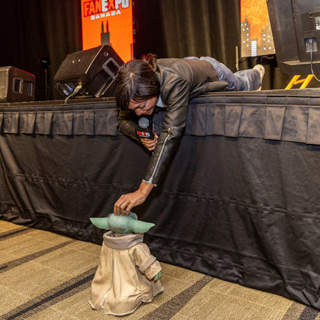 Rosario Dawson lays face down, hanging off the edge of the main stage. She pets an animatronic Grogu, which is facing her.
