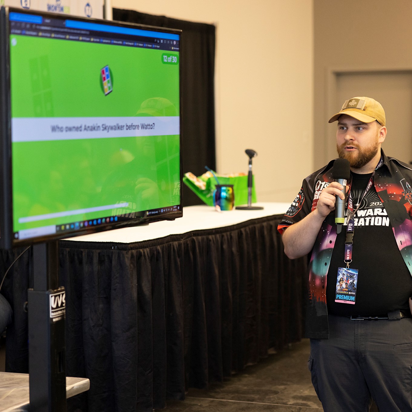 Screen with a green graphic with caption "Who owned Anakin Skywalker before Watto". Man standing on the right side while holding a microphone.