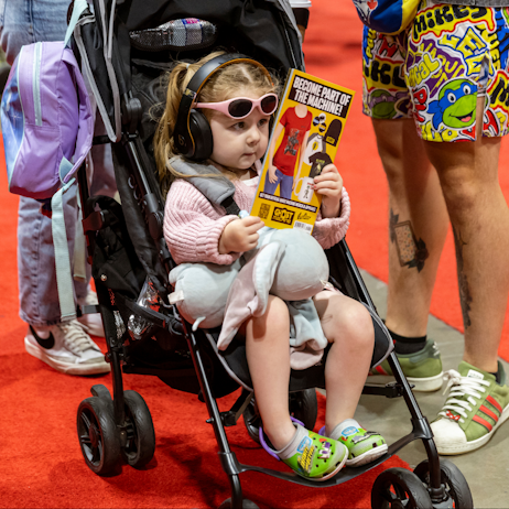 A little girl, wearing over ear headphones and pink sunglasses, holds a Ghost Machine flier up to her face to read it. She is in a stroller, with both parents standing on either side - only their legs are visible. 