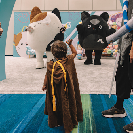A small child dressed as a jedi waves to two Squishmallow mascots. Left, is Cam, a brown, black, and white cat, and right is Emily, a black bat.