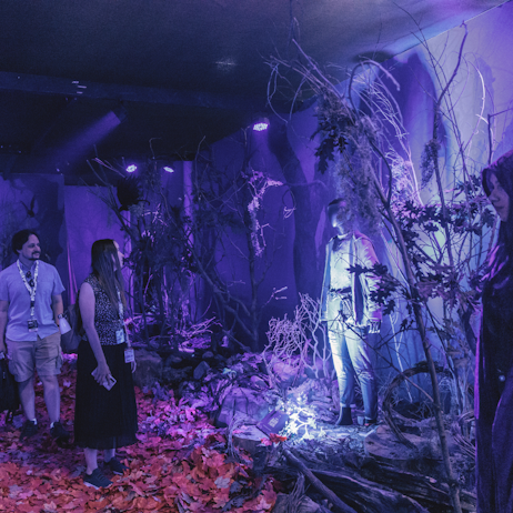 Fans walk the Witches' Road with Disney+ to see the costumes from Agatha All Along. The room is lit with purple lighting and dressed in trees and leaves to look like an old and decrepit forest.