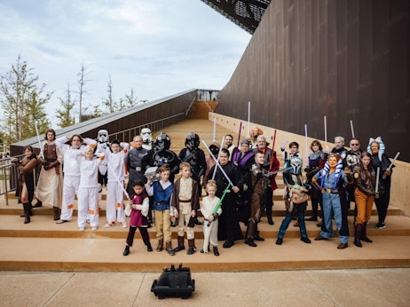 A diverse group of individuals, both adults and children, gather together outdoors, dressed in various "Star Wars" themed costumes. Characters range from Jedi Knights wielding lightsabers, Sith Lords, droids, and other iconic figures from the franchise. They stand proudly on a set of steps, with a modern architectural structure in the background, creating a blend of the fantasy world and contemporary surroundings. The atmosphere is celebratory, capturing the camaraderie of fans uniting in their shared passion