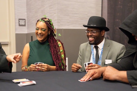 The image features two individuals engrossed in a joyful moment at a card game during a convention. On the left, a woman with striking maroon dreadlocks adorned with green leaves and wearing a forest green dress laughs heartily. To her right, a man in a classic gray suit, black tie, and a stylish hat shares the merriment, smiling broadly as he holds playing cards. The friendly and spirited interaction is set against a simple conference room backdrop, emphasizing the warmth and camaraderie of the scene.