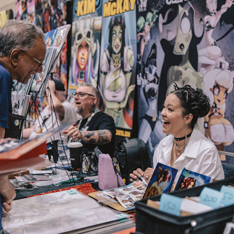 A fan interacts with a comic creator. Both parties are smiling wide as they chat. She is wearing little black cat ears in her dark hair. 