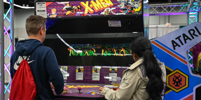Two individuals playing a retro arcade game called X-men. They both stand with backs to the camera.