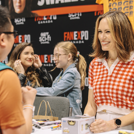 Emily Swallow smiles at a fan as he speaks to her. She holds their Armourer Funko Pop! in her hands. Beside her is a wide array of colour sharpies