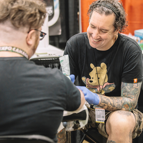 A tattoo artist works on a tattoo on a fan's forearm. He smiles as he works, and is wearing a tshirt featuring a fully tattooed Winnie the Pooh.