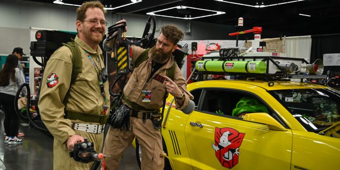 two individuals dressed in brown and green ghost busters jumpsuits, posing next to a yellow car with the ghostbusters logo on it.