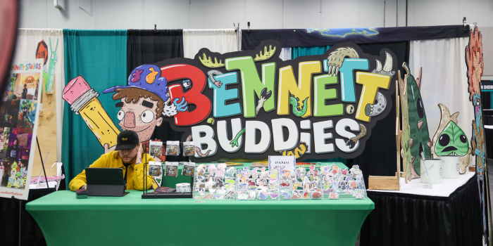 An image of the Bennett Buddies table with caricature designs and stickers on display. A man in a yellow jacket monitors the booth.