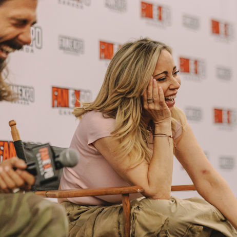 Tara Strong laughs during her panel at FAN EXPO Canada. She has a hand holding her chin and cheek as she leans on her knee. She is sitting in a director's chair in front of a white FAN EXPO Canada background. 