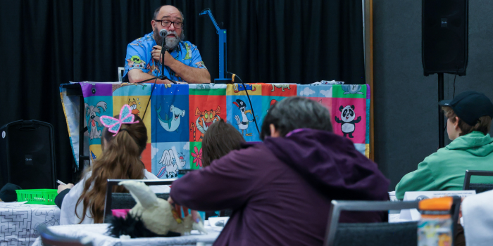 A man in a blue Hawaiian shirt sits at a table on a stage and teaches kids to draw cartoons. An audience of children sit around him drawing and listening.