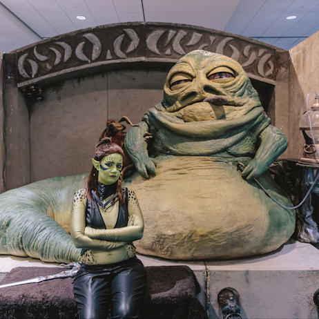 A Mirialan cosplayer poses, with a stern expression, with a large, faux Jabba the Hutt