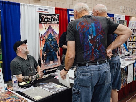 A man in a cap and grey shirt sits at a booth, laughing and looking up towards someone outside of the frame. He is behind a table displaying artworks and a banner that reads "Jeremy Clark". To the right, two men with their backs to the camera are looking at items on another table. One of them wears a t-shirt with a colorful Spider-Man design. The setting appears to be a comic or fan convention with various booths in the background.