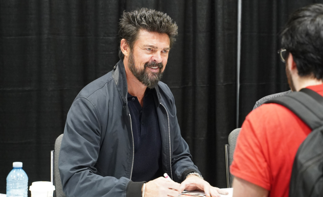 Karl Urban sits behind his autograph table as he signs a photo. He smiles at a fan whose back faces the camera.