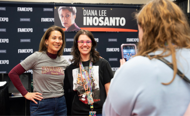 The image captures a moment at a FAN EXPO event, featuring two women posing for a photo with a fan. On the left, Diana Lee Inosanto smiles broadly; she is wearing a "Skywalker Ranch" graphic t-shirt over a long-sleeve burgundy top, paired with blue jeans. Beside her stands a delighted fan, wearing glasses and a black Star Wars-themed t-shirt adorned with a lanyard full of pins and badges. They are being photographed by another attendee, visible from behind. Behind them, a promotional banner featuring the name "Diana Lee Inosanto" adds to the atmosphere of the fan convention setting.