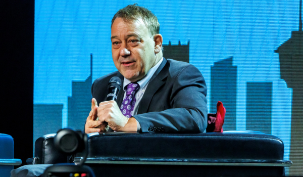 Sam Raimi is sitting in a black chair on stage and is holding a microphone. He is wearing a navy suit with a purple and white tie. He is speaking to a fan in the audience.