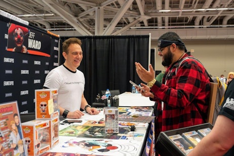 The image captures a cheerful interaction between Mike McFarland and an attendee at a FAN EXPO event. Mike, wearing a white t-shirt and smiling broadly, stands behind a booth adorned with various pop culture merchandise, including Funko Pops and colorful artwork. The attendee, dressed in a red and black plaid shirt and wearing a headband, is animatedly talking and gesturing with his hand, holding a smartphone in the other. The atmosphere is lively and engaging, highlighting the enjoyable exchanges that occur at such fan conventions.