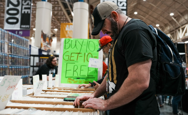 The image shows a man browsing items at a vendor's table in what appears to be a convention hall. He is wearing a black T-shirt, a cap, and has a backpack on, suggesting he is an attendee exploring the merchandise. The table is laid out with an array of comic books. Behind him, a sign reads "BUY 10 GET 10 FREE", indicating a special offer on the products. The vendor in the background seems engaged in a conversation with another person, possibly another attendee. The environment suggests a lively event where vendors and artists display their goods for sale, such as at a comic book or pop culture convention.