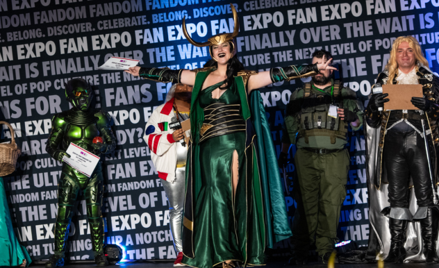 The image captures a moment on a stage at a cosplay event, with participants dressed in elaborate costumes. A central figure stands out, dressed in an elegant green gown with gold trim, arms outstretched in a theatrical pose that suggests a character of regality or power, possibly from a fantasy genre. To the left, the individual in the metallic green suit seen in a previous image is also present, holding a sign for the "COSPLAY CRAFTMANSHIP CUP." To the right of the central figure is a person dressed in military-style attire with a green vest, suggesting a character prepared for combat or survival. Further to the right, another individual in black and white with long blond hair seems to be portraying a historical or fantasy character.