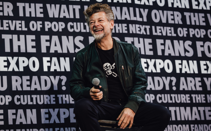 Andy Serkis with wavy gray hair and a beard sits in front of a backdrop with repeated text about a fan expo. He wears a black T-shirt with a skull graphic, a green leather jacket, and holds a microphone. He is smiling and seems to be engaged in a lively conversation.