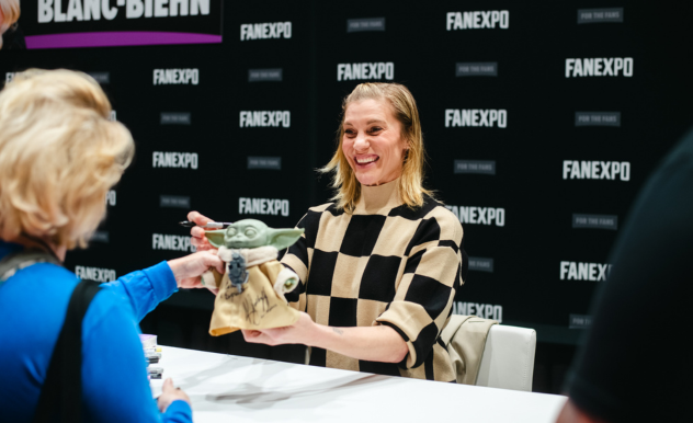The image captures a warm and cheerful interaction at a FAN EXPO event. Katee Sackhoff with a bright smile is sitting at a signing booth, adorned with the FAN EXPO branding, and is reaching out to receive a figurine of Yoda, presumably for an autograph, from a fan whose blue sleeve and blonde hair are visible in the foreground. The atmosphere appears to be light-hearted and engaging, indicative of the fan-friendly environment such conventions are known for. The woman’s striped top adds a casual and approachable vibe to the scene, suggesting she is a celebrity or artist who enjoys connecting with her audience.