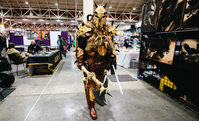 The image features an individual in an elaborate fantasy armor costume, walking through a convention hall. The armor is detailed and ornate, with bone and horn motifs, giving it a barbarian or warrior-like aesthetic. The costume includes a helmet with horns and appears to be made with a combination of materials to simulate metal, bone, and leather. The person is carrying a weapon prop that resembles a sword or large dagger. Vendor booths with various merchandise and artworks are visible in the background, and the scene suggests that this is a part of a larger event, likely a comic or fantasy convention, where cosplay is showcased.