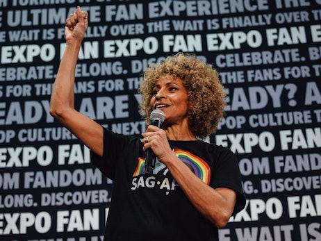 Michelle Hurd with curly golden-brown hair speaks into a microphone while on stage. She wears a black shirt featuring a rainbow design and raises one arm in a gesture of excitement. Behind her, a backdrop prominently displays repeated text, such as "FAN EXPO", "CELEBRATE", and "ARE YOU READY?", emphasizing the celebratory nature of the event. The woman's expression radiates passion and engagement with her audience.