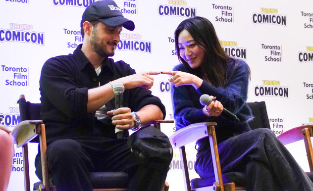 Tomer Capone (left) and Karen Fukuhara (Right) are seated in director's chairs. They lean in towards each other with index fingers extended, touching the tips of their fingers