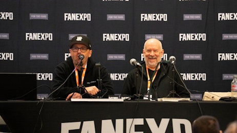 The image depicts two men sitting at a panel during a convention, both engaging with an audience. The man on the left is wearing glasses and a black cap, focused intently on speaking into a microphone. Beside him, the other man, bald and with a goatee, wears a warm smile and an orange lanyard, listening attentively to his colleague. They are seated behind a table with a FAN EXPO banner in the background, enhancing the professional and interactive atmosphere of the event.
