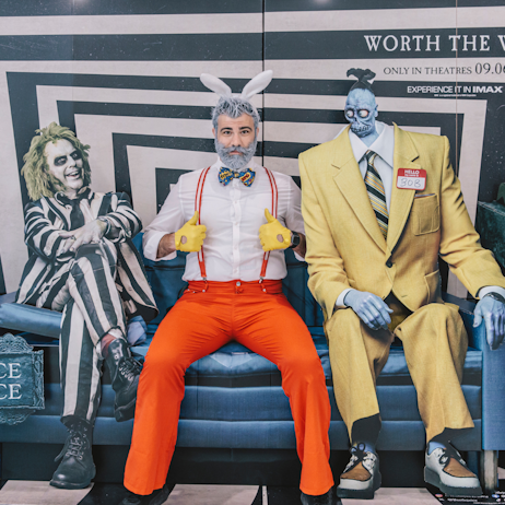 A fan dressed as Roger Rabbit, who has added white spray-in dye to their hair and beard, sits between Beetlejuice and "BOB" the Shrinker, on a blue cardboard couch. This standee is a sponsorship ad for Warner Brothers and the release of Beetlejuice Beetlejuice