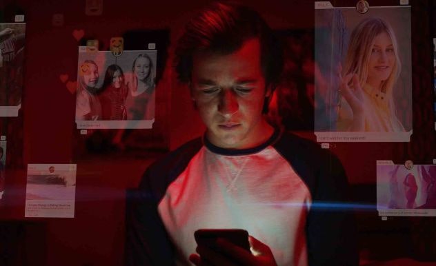 A teenager looks at his phone alone in a dark room with images of the social media posts he's looking at floating translucently around him
