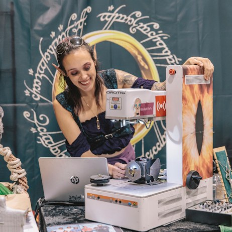 A person wearing elf ears and Elven garb uses an engraver (with Sauron's eye printed on it) to engrave a piece of custom jewelry for a fan.