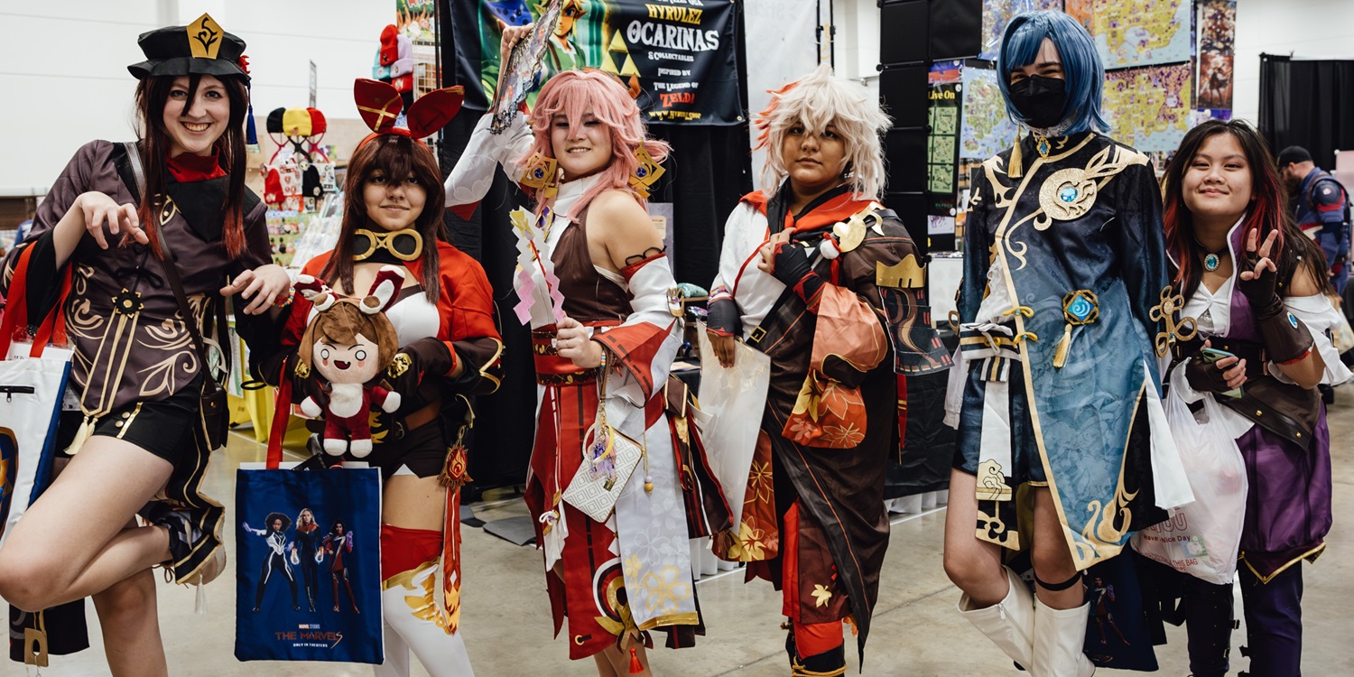 A group of cosplayers dressed as characters from Genshin Impact poses together at FAN EXPO. Each cosplayer displays their intricate costumes, accessories, and props, capturing the vibrant spirit of the game. They stand in front of a colorful booth, smiling and holding bags with merchandise from the event.
