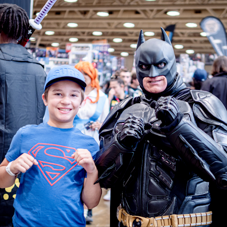 A young fan in a Superman tshirt poses with a Batman cosplayer on the expo floor. They are holding their fists up as if they are ready to throw a punch, and smiling.