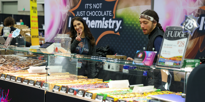 Two vendors smile for camera and stand at their fudge display. Display shows an arrangement of colorful fudge options for purchase.
