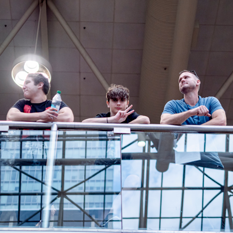 Fans lean on the railing above the North building escalators. While two look off into the distance, the youngest of the three is looking down, posing with a peace sign for the camera. 