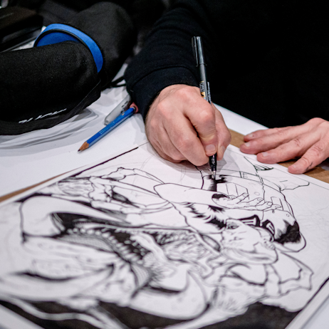 A comic artists' hands use a black felt pen to draw a new image. In the image, a creature appears to be chasing an unsuspecting victim who is human and male presenting.
