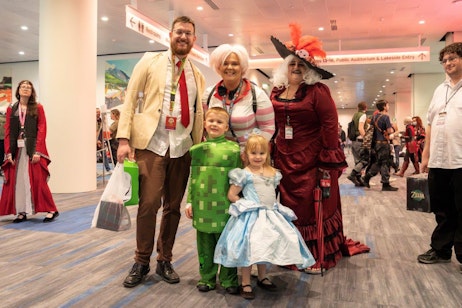 The image features a group of six people in vibrant cosplay at a convention. On the left, a man in a light jacket and tie represents a fandom, while a woman next to him wears an elaborate red and black costume. Centered are two young children: one dressed as a green pixelated character from a video game and the other as a classic fairy tale princess in a blue gown. On the right, a woman in a large red dress with a dramatic hat poses playfully alongside another person in casual attire carrying a book. The setting includes a bustling convention hall with signage pointing to facilities and event areas.