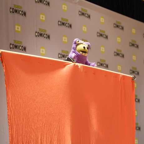 Mona from Nanalan, the small green puppet, pops over an orange sheet that hides her puppeteer. Mona is wearing a purple monster costume.