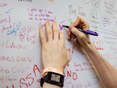 A close-up of a hand holding a purple marker, writing on a white board filled with various colorful handwritten messages and drawings. Some notable messages include "7 years ago I came here on a first date... and he's now my husband!" and "The Force is...". The board is a mix of personal anecdotes, shout-outs, and playful doodles, showing the collective engagement of multiple individuals. The writer wears a watch on their wrist.